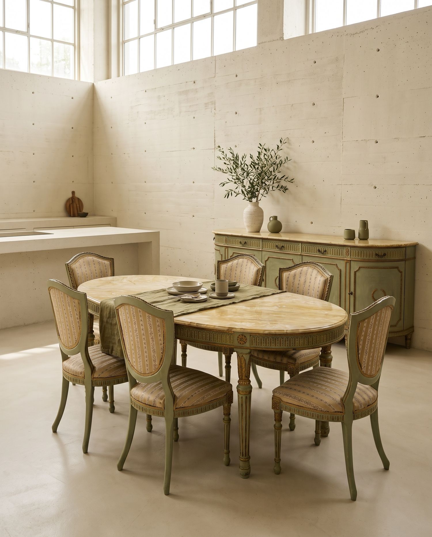 Tavolo ovale con piano in marmo giallo e sei sedie dipinte in verde salvia, con la credenza abbinata contro una parete in cemento chiaro